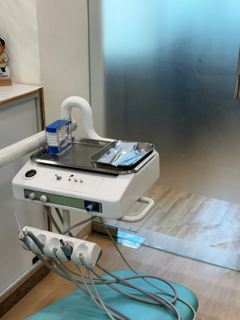 This image displays a complete dental treatment setup inside a modern clinic. An orange reclining dental chair is positioned centrally, accompanied by an overhead examination light, instrument console, and mounted display screen. Nearby cabinetry and dental tools are organized for easy access. The room has large windows and frosted glass partitions, creating a bright, clean, and professional clinical environment suitable for patient consultations and procedures.